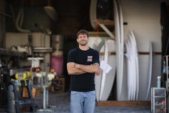 Person standing in a workshop with surfboards, tools, and equipment visible in the background.